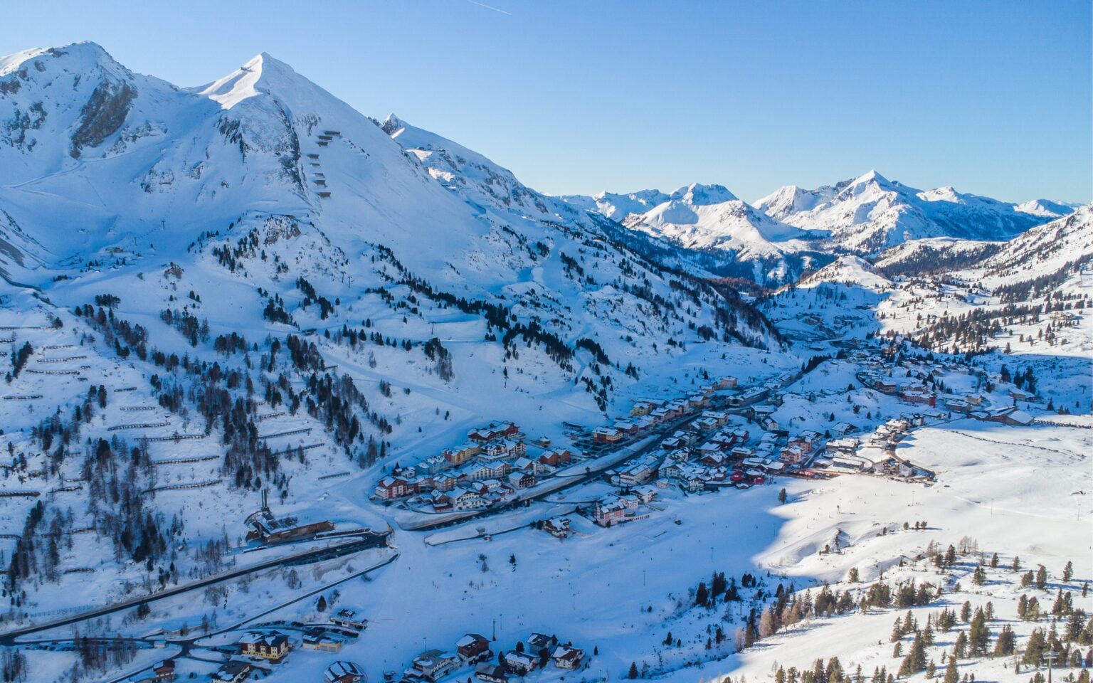 Der Skiort Obertauern mit Gebäuden, Nadelbäumen und der umliegenden Bergwelt an einem sonnigen Tag.