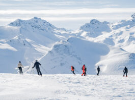 Eine Gruppe von Menschen, die auf Skiern oder Snowboard eine Skipiste in den Alpen hinunterfahren.