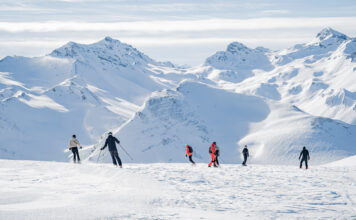 Eine Gruppe von Menschen, die auf Skiern oder Snowboard eine Skipiste in den Alpen hinunterfahren.