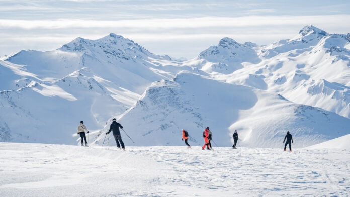 Eine Gruppe von Menschen, die auf Skiern oder Snowboard eine Skipiste in den Alpen hinunterfahren.