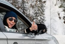 Lächelnde Frau sitzt in einem Auto und zeigt den Daumen nach oben. Sie ist auf einer verschneiten Straße unterwegs.