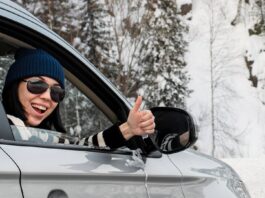 Lächelnde Frau sitzt in einem Auto und zeigt den Daumen nach oben. Sie ist auf einer verschneiten Straße unterwegs.