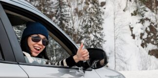 Lächelnde Frau sitzt in einem Auto und zeigt den Daumen nach oben. Sie ist auf einer verschneiten Straße unterwegs.