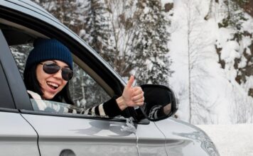 Lächelnde Frau sitzt in einem Auto und zeigt den Daumen nach oben. Sie ist auf einer verschneiten Straße unterwegs.