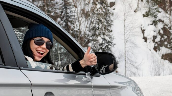 Lächelnde Frau sitzt in einem Auto und zeigt den Daumen nach oben. Sie ist auf einer verschneiten Straße unterwegs.