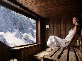 Eine schöne Frau mit einem weißen Handtuch geht in die Sauna. Die Sauna ist aus Holz und hat ein großes Fenster mit Blick auf die Berge.