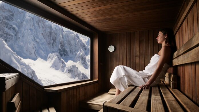 Eine schöne Frau mit einem weißen Handtuch geht in die Sauna. Die Sauna ist aus Holz und hat ein großes Fenster mit Blick auf die Berge.