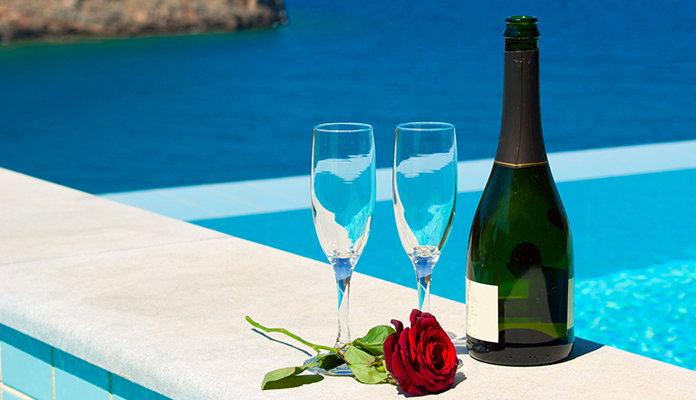 Två glas står bredvid en flaska som liknar bubbel, med en ros intill och utsikt över en pool och havet i bakgrunden.